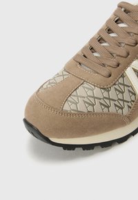 Sneaker en suède marron clair avec un motif géométrique beige, des lacets gris et une semelle en caoutchouc noir texturé.
