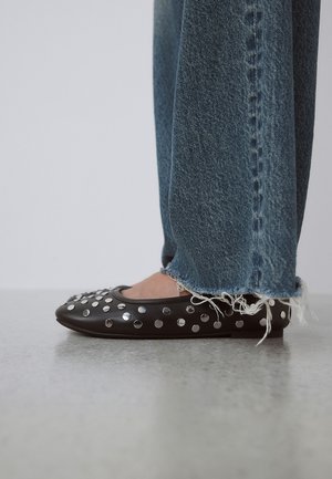 Fekete lapos cipő ezüst szegecsekkel, kék rojtos szegélyű farmerral, szürke padlón.