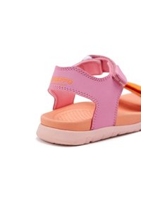 Sandalia rosa con correa ajustable y acento naranja. Presenta una suela texturizada y materiales suaves. Incluye un logo en naranja en el talón.
