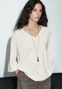 Cream v-neck sweater with relaxed fit and 3/4 sleeves, paired with taupe textured pants. Features layered gold necklaces.