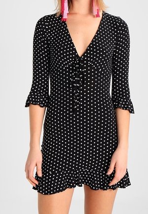 Sort kjole med hvide prikker med 3/4-ærmer, flæsekant ved ærmer og nederdel, dyb V-udskæring og bindebånd foran, båret med lange, lyserøde kvastøreringe.