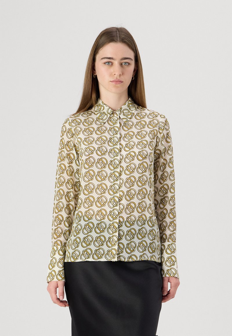 Camicia a maniche lunghe con bottoni realizzata in un tessuto leggero. Presenta una stampa geometrica ripetitiva in verde e marrone su sfondo crema.