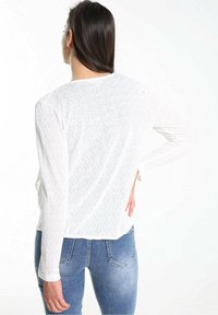 Haut blanc à manches longues avec un motif texturé, doté d'un col rond et d fentes sur les côtés, associé à un jean en denim bleu.