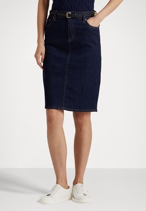 Kvinde iført en mørkeblå denim nederdel til knæene med sort bælte, hvide sneakers og en mørkeblå top med den ene hånd i lommen.