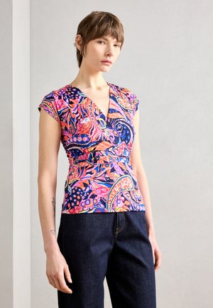 Haut ajusté et coloré avec un décolleté en V plongeant, des manches courtes et un motif paisley vibrant sur un fond sombre. Associé à un jean foncé.