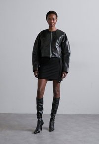 Fekete, fényes bőr bombázó dzseki cipzárral, fekete mini ruhára és térd fölé érő fekete magassarkú csizmára húzva.