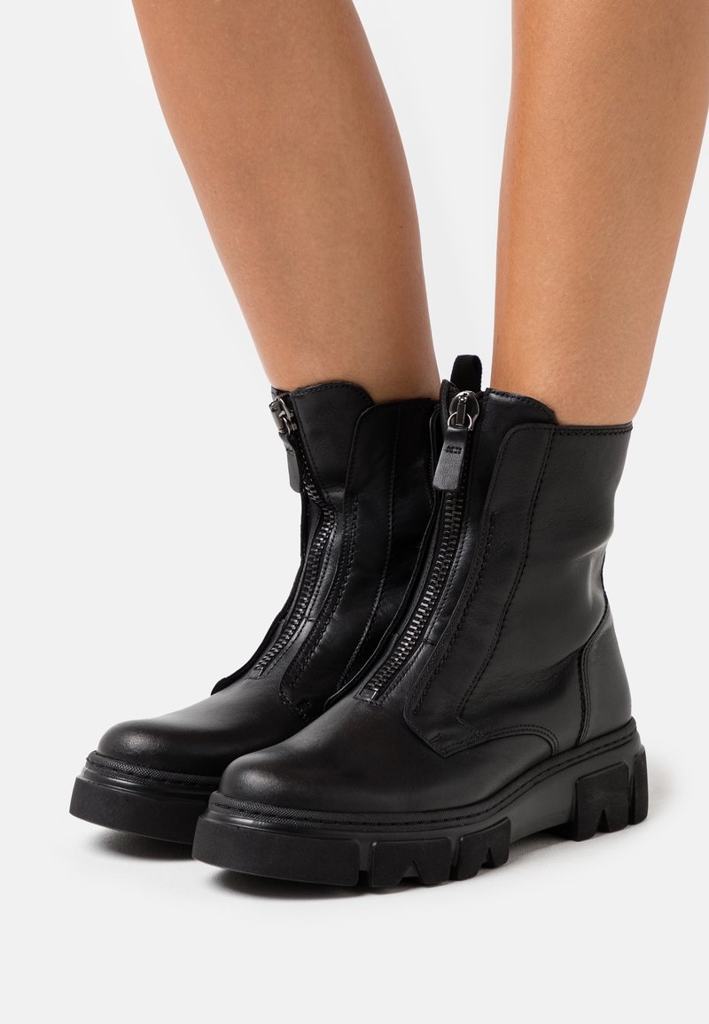 Jambes portant des bottes mi-mollet en cuir noir avec des semelles épaisses et des fermetures éclair verticales à l'avant, debout sur une surface blanche.