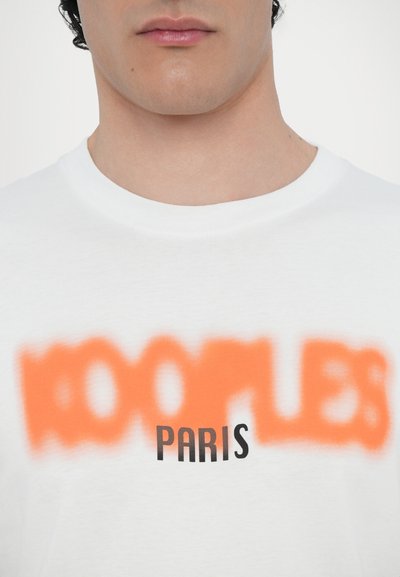 T-shirt en coton blanc avec un texte orange flou "KOOPLES" au-dessus d'un "PARIS" noir clair, col rond et texture lisse.
