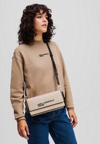 Borsa a tracolla beige con rifiniture nere e logo, con una texture liscia simile alla pelle e una tracolla staccabile, indossata con una felpa coordinata.