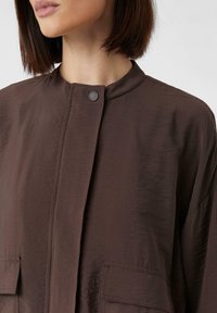 Braune, leichte Jacke mit einem hohen Kragen, zwei Fronttaschen und einem Druckknopfverschluss, aus einem glatten, leicht strukturierten Stoff.