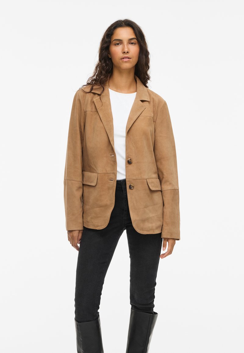 Tan suede blazer with notched lapels, two front pockets, and two buttons. Worn over a white top with black skinny jeans and tall black boots.