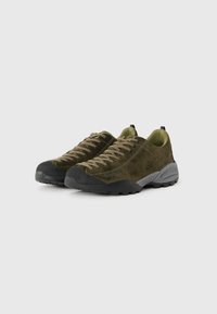 Une paire de chaussures de randonnée en suede vert olive avec des semelles en caoutchouc noir et des lacets beige, présentée sous un léger angle sur un fond uni.