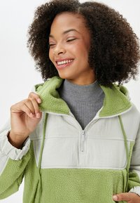 Sudadera de forro polar en verde claro y gris con cuello de media cremallera, que presenta un acabado texturizado y un diseño de bloques de color.