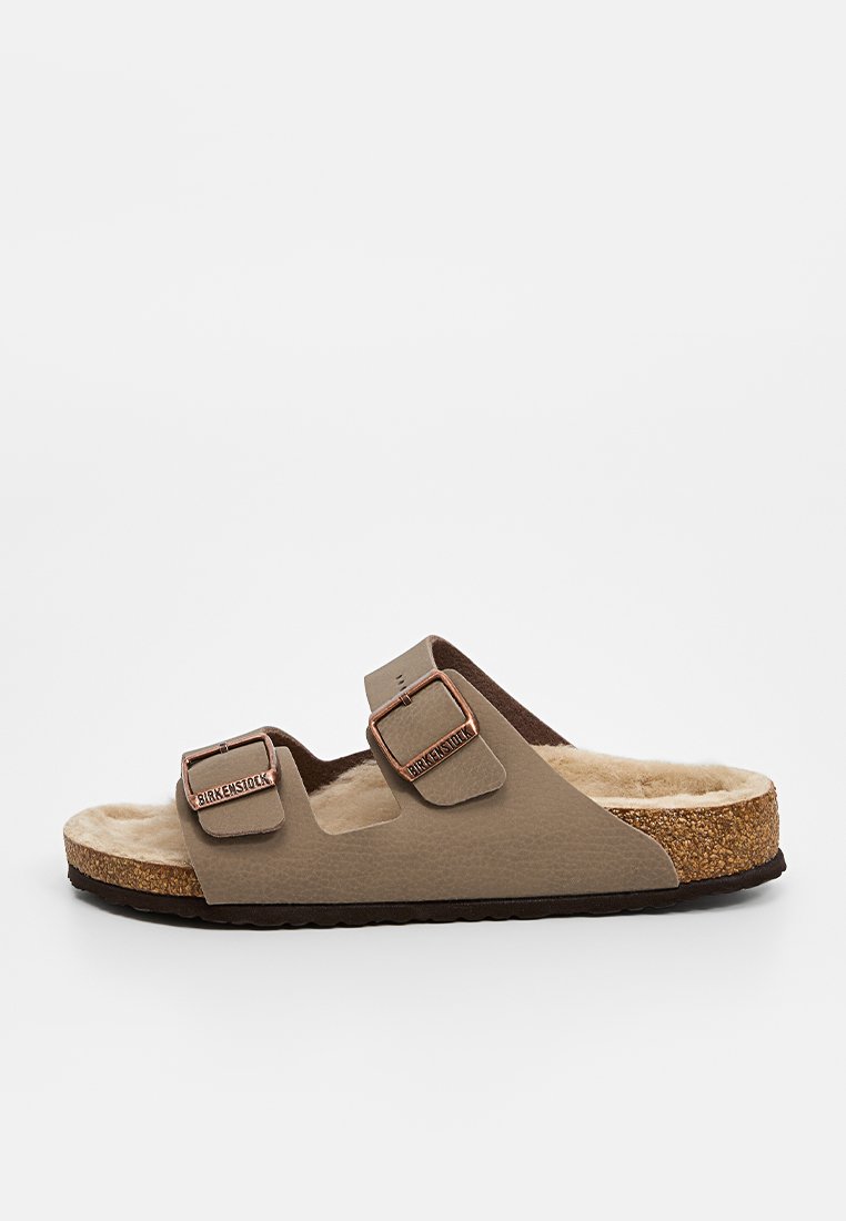 Sandale brune à deux sangles avec des boucles, dotée d'une semelle intérieure en fourrure douce et d'une semelle extérieure en liège. Fabriquée en matériau synthétique avec une texture lisse.