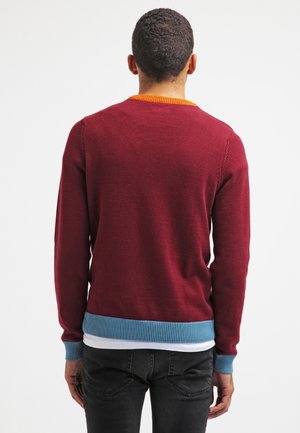 Homme vu de dos portant un pull bordeaux avec des poignets et un ourlet bleus, un col orange, et un jean noir sur un fond blanc uni.