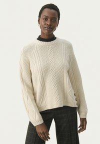 Cremefarvet strikket sweater med et tekstureret design med kabler og mønstre, ribbede kanter og knapper i siderne. Båret med mørke mønstrede bukser.