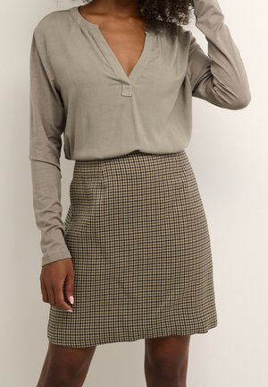 Kvinde iført en taupe langærmet V-hals top, stoppet ned i et højtaljet brun og sort hundetand mønsteret skørt mod en ensfarvet baggrund.