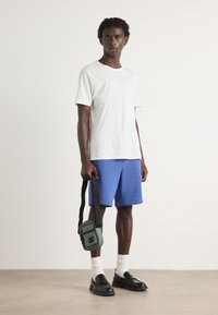 Tricou alb cu mânecă scurtă, pantaloni scurți albaștri, mocasini negri, șosete albe, ținând o geantă mică verde tip crossbody cu un logo negru și roșu.