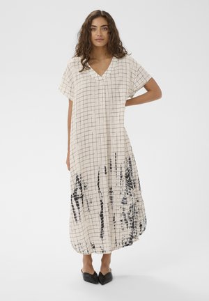 Vrouw met lang krullend haar draagt een losse, tot aan de enkels vallende crèmekleurige jurk met een zwart raster- en abstract patroon, gecombineerd met zwarte puntige schoenen.