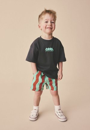 Noor poiss naeratab, seljas must T-särk kirjaga "COOL", roheliste ja punaste triipudega lühikesed püksid, valged sokid ja valged takjakinnitusega tossud, seisab beeži tausta ees.