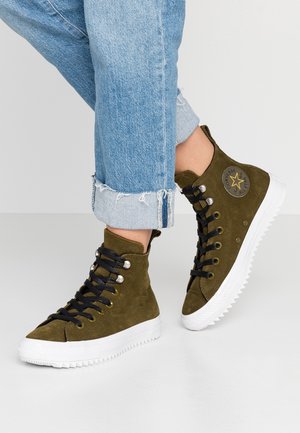 Person iført opkrogede blå jeans og olivengrønne høje Converse-sneakers med sorte snørebånd og hvide robuste såler.