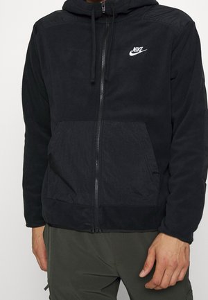 Person trägt einen schwarzen Nike-Kapuzenpullover mit durchgehendem Reißverschluss, Fronttasche und verstellbarer Kapuze, kombiniert mit dunkelgrünen Hosen.