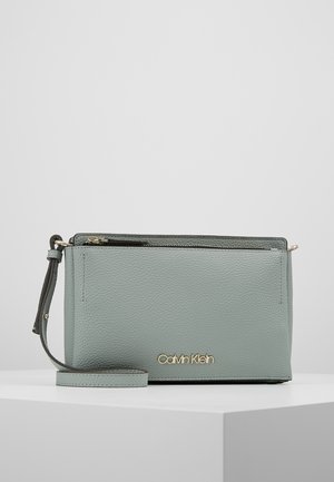 Petit porte-monnaie bracelet en cuir gris clair Calvin Klein avec logo doré, fermeture à glissière et sangle de poignet amovible, présenté sur un socle blanc.