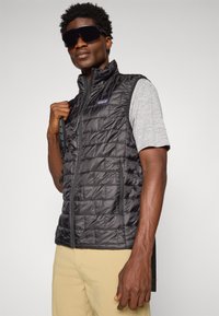 Black puffy vest with zip closure, horizontal quilting, and gray fabric sleeves. Notable details include side pockets and a logo.
