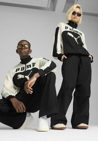 Black and white tracksuits with a prominent Puma logo, paired with sunglasses. Loose-fitting design and textured fabric. Casual footwear.