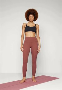 Μαύρο σουτιέν αθλητικής ένδυσης με λεπτές τιράντες, ψηλόμεσα κολάν σε πορτοκαλί απόχρωση, και τα δύο κατασκευασμένα από ελαστικό ύφασμα, πάνω σε μια ροζ στρώμα γιόγκα.