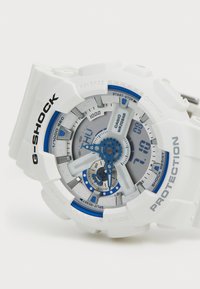 Montre G-Shock avec un boîtier et un bracelet en résine blanche, accents bleus, affichage rond analogique-numérique, boutons texturés et caractéristiques de résistance aux chocs.