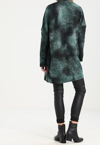 Cappotto lungo a macchie verdi e nere con una vestibilità rilassata, abbinato a leggings in pelle nera e stivaletti. Texture morbida e design drappeggiato.