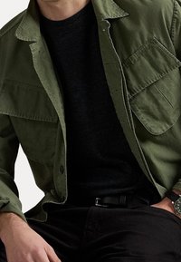 Man som bär en öppen grön skjorta med knappar över en svart t-shirt, svarta byxor med bälte, och en grön armbandsur på v änster handled.