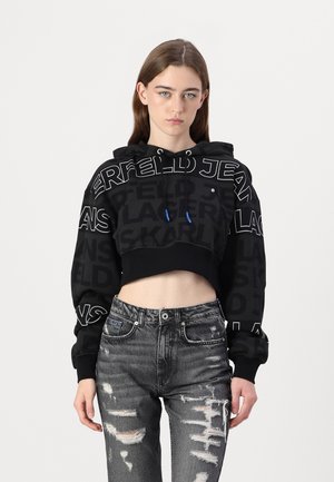 Felpa nera cropped con testo bianco audace e lacci blu, abbinata a jeans grigi strappati con strappi e una vestibilità affusolata.