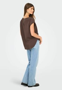 Donna che indossa una maglietta marrone a maniche corte con zip sul retro, jeans a gamba larga blu chiaro, stivali neri e orecchini argentati, in piedi di profilo.