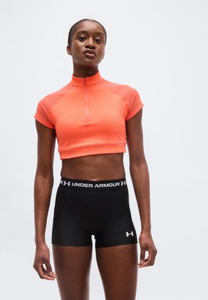 Nő narancssárga, rövid, cipzáras felsőben és fekete Under Armour sportnadrágban, egy egyszínű fehér háttér előtt áll.