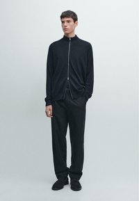 Schwarze Zip-Up-Strickjacke mit hohem Kragen, kombiniert mit locker sitzenden dunklen Hosen und schwarzen Schuhen, vor hellem Hintergrund.