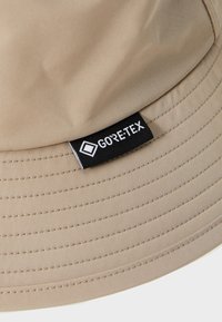 Cappello a secchiello in GORE-TEX beige con una finitura liscia e resistente all'acqua. Presenta dettagli cuciti e un'etichetta con logo nera sul lato.
