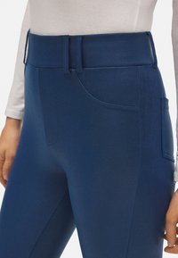 Pantalones azul marino con una textura suave, cintura alta, pasadores para cinturón y un bolsillo lateral. Presentan un diseño ajustado y detalles de costura sutiles.