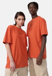 Afslappede orange bomulds t-shirts med korte ærmer, der har et diskret sort signaturtryk på brystet, parret med beige cargo bukser.