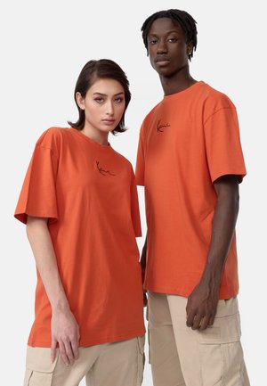 Afslappede orange bomulds t-shirts med korte ærmer, der har et diskret sort signaturtryk på brystet, parret med beige cargo bukser.