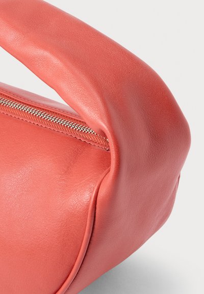 Κοντινή λήψη ενός τσάντας χρώματος κοραλλί από δέρμα με καμπυλωτή λαβή και εμφανές ασημένιο φερμουάρ σε ανοιχτό φόντο.