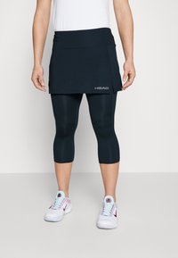 Marinblå sportkjol med platt midja, med en sidoklyvning och varumärkets logotyp. I kombination med matchande långa leggings och vita sneakers.