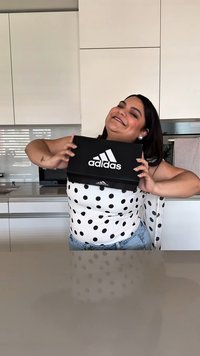 Caixa preta da Adidas com o logótipo branco, segurada por uma pessoa com uma blusa de bolinhas. O fundo da cozinha moderna apresenta armários e bancadas elegantes.