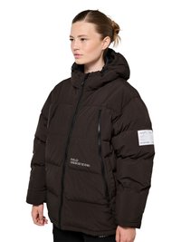 Kobieta ubrana w czarną pikowaną kurtkę z kapturem, zamkami błyskawicznymi i białym napisem z przodu oraz na lewym rękawie, stojąca na białym tle.