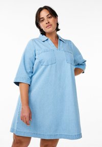 Šviesiai mėlynas džinsinis suknelė su apykakle, trumpomis rankovėmis, dviem krūtinės kišenėmis ir laisvu siluetu, turinti v formos iškirpę ir sijoną, kuris baigiasi virš kelių.