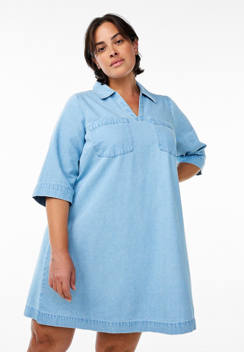 Šviesiai mėlynas džinsinis suknelė su apykakle, trumpomis rankovėmis, dviem krūtinės kišenėmis ir laisvu siluetu, turinti v formos iškirpę ir sijoną, kuris baigiasi virš kelių.