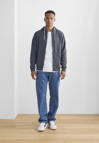 Sweat à capuche gris zippé avec cordons, superposé à un t-shirt blanc, associé à un jean en denim bleu et des baskets blanches. Tissu lisse, coupe décontractée.