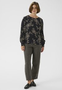 Moteris stovi vilkėdama juodą gėlių rašto ilgą rankovėmis bluzoną, pilkas tiesaus kirpimo kelnes ir juodus loaferius prieš vienodą foną.