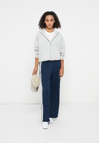 Gray zip-up hoodie with long sleeves, paired with navy wide-leg pants featuring white side stripes. Holding a beige cap, white sneakers visible.